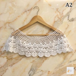 May include: A white, open-weave crochet collar with a scalloped edge, displayed on a wooden hanger. The collar has a delicate, lacy pattern and is against a marble-patterned background. The text "A2" is visible in the upper right corner.