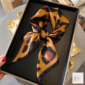 May include: A silk scarf with a leopard print design. The scarf has a tan background with black and brown spots. The scarf is displayed on a black tray with a gold frame.