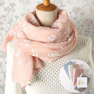 May include: A soft peach-colored scarf featuring a white floral pattern, draped over a mannequin. The scarf has a lightweight, semi-sheer texture. A small circular display shows four folded scarves in blue, cream, peach, and gray with floral designs.