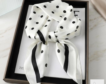 May include: White silk scarf with black polka dots and vertical black stripes. The scarf is tied in a knot and displayed in a dark brown tray. The scarf is made of a soft, flowing material.