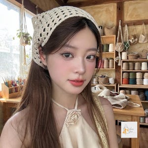 May include: A cream-colored crochet headscarf with a diamond-shaped openwork pattern. The headscarf is worn over long brown hair and is paired with a matching cream-colored halter top featuring a fabric rose detail at the center of the neckline.