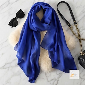 May include: A vibrant royal blue scarf with a smooth, semi-sheer texture. The scarf is draped over a cream-coloured faux fur piece, accompanied by black sunglasses with dark lenses and a black handbag featuring a gold-tone chain shoulder strap.