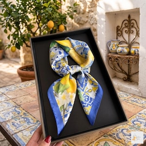Puede incluir: Un pañuelo de seda con un diseño de limón y flores, presentado en una bandeja negra. El pañuelo presenta un borde azul y una mezcla de patrones amarillos, blancos y azules. El fondo incluye un limonero y cerámica decorativa.