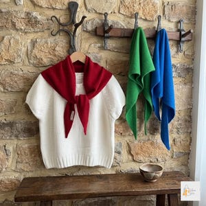 Puede incluir: Un suéter de punto blanco de manga corta en una percha de madera con una bufanda roja atada al cuello. Dos bufandas adicionales en verde y azul cuelgan de un perchero de pared junto al suéter. Un pequeño cuenco de cerámica descansa en un banco.
