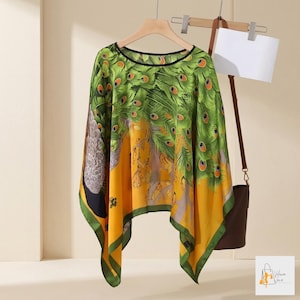May include: A lightweight poncho featuring a vibrant peacock feather print in shades of green, yellow, and black. The garment has a wide, draped silhouette with a rounded neckline and a black trim along the edges, hanging from a wooden hanger.