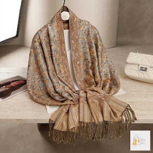 May include: A patterned scarf in gold, brown, and grey, with a fringed edge. The scarf is draped over a white top on a hanger. A white handbag is in the background.