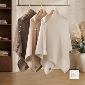 May include: Three soft knit ponchos hanging on a gold metal rack. The ponchos are in shades of dark brown, light beige, and cream. Each garment features a vertical row of small, round, metallic buttons along one side and a small rectangular tag.