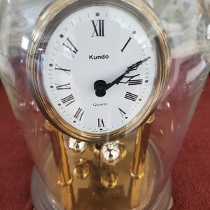 May include: A gold-toned Kundo quartz clock with Roman numeral markings on the face, encased in a clear glass dome. The clock is on a wooden base with a Kundo Quartz logo.