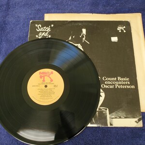 May include: A black vinyl record with a gold and pink label, titled "Satch and" and "Count Basie encounters Oscar Peterson." The album cover features a black and white image of a man smoking a cigarette.