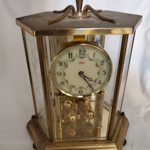 May include: A gold-toned, octagonal, glass-enclosed, mechanical clock with a white face and black hands. The clock face has a red floral design and the word "Almada" printed on it.