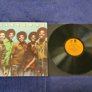 May include: A vinyl record album cover featuring a group of five men, The Jacksons, with the album title "The Jacksons" printed on the cover. The album cover is in a blue and green color scheme. The record is black.