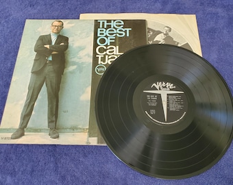 Può includere: Un disco in vinile nero con un'etichetta bianca che dice "The Best of Cal Tjader" e "Verve Records". Il disco è su uno sfondo bianco con una foto di un uomo in abito.