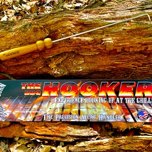 Könnte beinhalten: Der BBQ Hooker, ein Edelstahl-Fleischhaken mit einem Holzgriff, ist auf einem Baumstamm abgebildet, zusammen mit einer schwarzen und roten Verpackung, auf der "The BBQ Hooker - Experience hooking up at the grill - The Precision Meat Handler - Certified" steht.