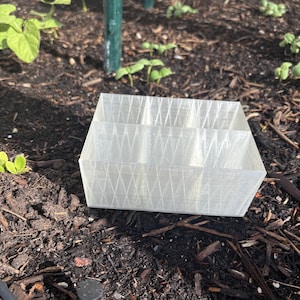 Puede incluir: Un divisor de lecho de jardín rectangular de plástico translúcido con múltiples compartimentos. El divisor se coloca en un lecho de jardín con tierra oscura y plantas jóvenes. El divisor tiene un patrón entrecruzado.