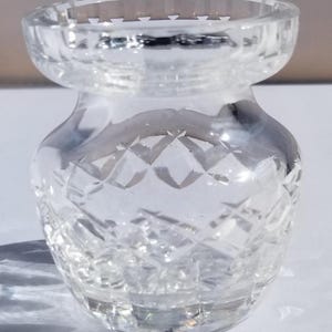 May include: A clear, small crystal vase with a diamond-cut pattern. The vase has a rounded base, a narrow neck, and a flared rim. The glass is transparent, allowing light to pass through, creating shadows. The vase is likely used for displaying flowers or as a decorative item.