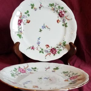 May include: Two white porcelain plates with scalloped edges and floral designs. The plates feature a gold rim and are decorated with colorful flowers, including pink, red, and blue blooms. The plates are displayed on a dark wooden stand against a burgundy background.