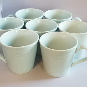 May include: A set of seven light green ceramic mugs. Each mug has a cylindrical shape with a handle and a textured, ridged exterior. The mugs are arranged in a group, showcasing their similar design and color.