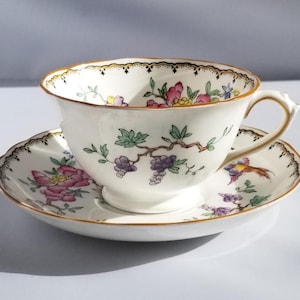 May include: A white teacup and saucer set with a floral design. The cup and saucer have a gold trim and a scalloped edge. The floral design includes pink, purple, and green elements.