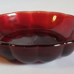 May include: A ruby red glass dish with a scalloped edge and a handle. The dish is shaped like a leaf and has a smooth, glossy surface. The handle has a stepped design. This decorative dish is suitable for serving small portions.
