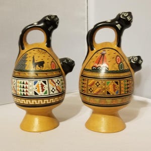 May include: Two ceramic jugs with black animal figures on the handles. The jugs are decorated with geometric patterns in black, red, green, yellow, and blue. The jugs have a light brown base.