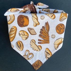 May include: White cotton bandana with a brown and white pattern of various breads and pastries. The bandana is tied in a knot at the top.