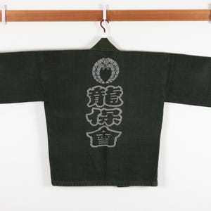 以下が含まれることがあります： 背中が白く刺繍された、濃い緑色の日本のジャケット。デザインは、花が描かれた円形の中に、その下に縦書きで「龍虎」の文字が書かれている。
