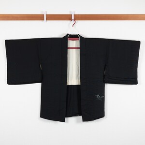 May include: A black kimono with a white lining and a small embroidered design on the right side. The kimono is hanging on a wooden hanger.