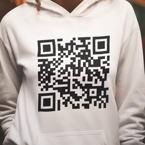May include: A woman wearing a white hoodie with a black QR code printed on the front. She is smiling and looking up.