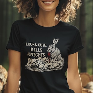 May include: Black t-shirt with the text "LOOKS CUTE. KILLS KNIGHTS" and a graphic of a rabbit sitting on skulls. The person is wearing khaki pants. The background is a forest.