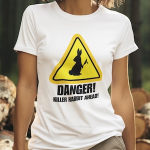 Könnte beinhalten: Weißes T-Shirt mit einem gelben Warnschild mit einem schwarzen Kaninchen, das eine Waffe hält. Der Text "DANGER! KILLER RABBIT AHEAD!" ist darunter gedruckt. Die Person trägt eine khakifarbene Hose und steht in einem Wald.