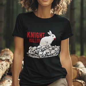 Peut inclure: T-shirt noir avec l'inscription "KNIGHT KILLER" en rouge et blanc. Le motif représente un lapin blanc au nez rouge sur un tas de crânes. La personne porte un pantalon kaki.