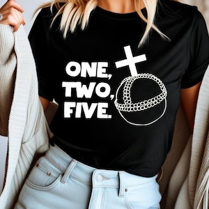 Könnte beinhalten: Schwarzes T-Shirt mit den Worten "ONE, TWO, FIVE" in fetten weißen Buchstaben. Ein weißes Kreuz und ein Perlenkreis-Design sind ebenfalls abgebildet. Das Shirt hat einen klassischen Rundhalsausschnitt.