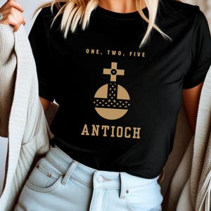 May include: Black t-shirt with a gold graphic featuring a cross atop a globe design and the word "ANTIOCH" below. The shirt also has the text "ONE, TWO, FIVE" above the graphic. The shirt is being worn.