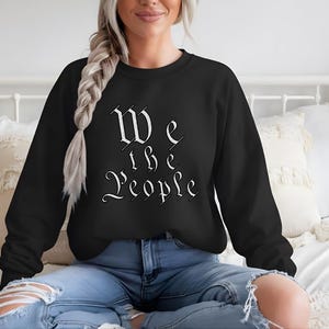 Op de afbeelding: Een zwart sweatshirt met lange mouwen en een ronde hals, voorzien van de witte tekst We the People in een gestileerd gotisch lettertype op de borst. Het kledingstuk heeft een geribbelde halslijn, boorden en zoom voor een standaard pasvorm.
