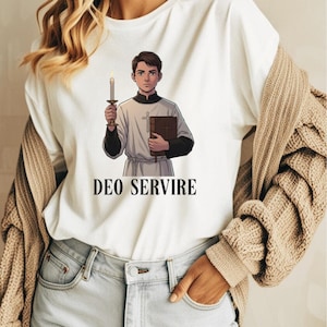 May include: A white t-shirt featuring a graphic illustration of a young person in a white and black liturgical garment holding a lit candle and a brown book, with the text DEO SERVIRE printed in black capital letters underneath the illustration.