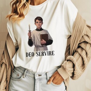 May include: White t-shirt with a cartoon illustration of a person holding a candle and a book. The text "DEO SERVIRE" is printed in black below the illustration. The person is wearing a white robe and black sleeves.