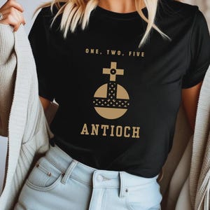 Puede incluir: Una camiseta negra de manga corta con un gráfico dorado de un orbe rematado con una cruz. Sobre el orbe, el texto dice ONE, TWO, FIVE, y debajo del orbe, el texto dice ANTIOCH. La camiseta tiene cuello redondo y un corte estándar.