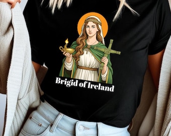 Puede incluir: Camiseta negra con un diseño de Brígida de Irlanda, una mujer con un halo, sosteniendo una lámpara y una cruz. El diseño incluye el texto "Brigid of Ireland" en blanco. La mujer lleva un vestido blanco, una capa verde y una corona dorada.