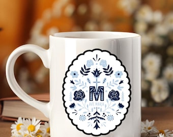 May include: White ceramic mug with a decorative oval design featuring blue floral elements and the text "AVE MARIA". The design includes roses, leaves, and the letter "M" with a cross. The mug has a curved handle.