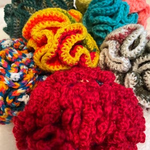May include: A collection of handmade crochet scrunchies in various colors. The scrunchies feature a textured, ruffled design. Colors include red, yellow, orange, blue, green, and a multi-colored scrunchie with white, blue, and orange.