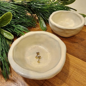 May include: Two white ceramic bowls, one slightly larger than the other, with a rough, textured surface. The smaller bowl contains two gold-coloured earrings.