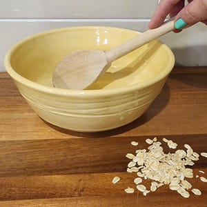 May include: A pale yellow ceramic mixing bowl with a wooden spoon inside. Rolled oats are scattered on the wooden surface. The bowl has a ridged design around the exterior. A hand with a teal nail holds the spoon.