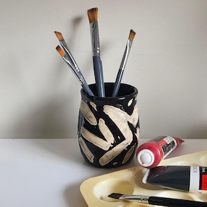 May include: A black ceramic paintbrush holder with gold abstract designs. The holder is filled with four paintbrushes. A red and black paint tube is visible in the foreground.