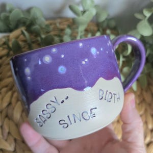 May include: A ceramic mug with a purple upper section featuring white speckled details and a cream-coloured lower section. The text SASSY.. SINCE BIRTH is embossed in dark lettering across the cream portion. The mug has a curved handle.