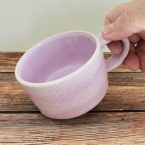 May include: A lilac ceramic mug with a handle. The mug has a speckled texture and a white rim. The mug is held by a hand and is set against a wooden surface.