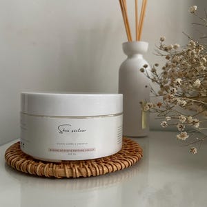 Op de afbeelding: Een witte pot Shea Senteur body butter staat op een ronde, geweven onderzetter. De pot heeft een etiket met Franse tekst. Een witte diffuser met riet en droogbloemen staat op de achtergrond. De body butter is 200 ml.
