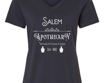 Può includere: Una maglietta blu scuro a collo a V con un design grafico bianco che recita "Salem Apothecary" con il testo "Tinctures, Potions & Elixirs" e "Est 1692" sotto.