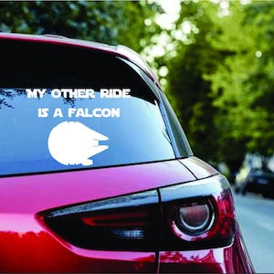 Puede incluir: Un coche rojo con una calcomanía de vinilo blanco en la ventana trasera. La calcomanía dice "My other ride is a Falcon" y presenta una silueta de la nave espacial Millennium Falcon.