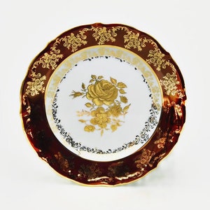 Może przedstawiać: Biały talerz porcelanowy z złotym wzorem kwiatowym i bordową obwódką. Talerz ma falisty brzeg i jest ozdobiony złotą obwódką.