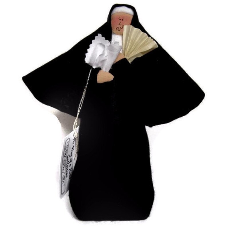 May include: A black fabric nun doll with a white head and a white fan. The doll is holding a white bouquet of flowers. The doll is wearing a black habit and a white veil. The doll is labeled "The Nun of a Kind Collection, Sweet Sister Grace".